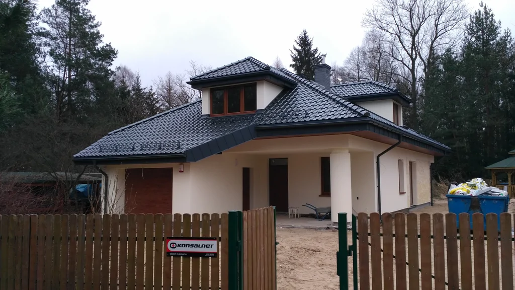 Jasna elewacja strefa wejścia, dach pokryty blachą na rąbek, okrągły słup, brązowa stolarka okien, komin obmurowany klinkierem