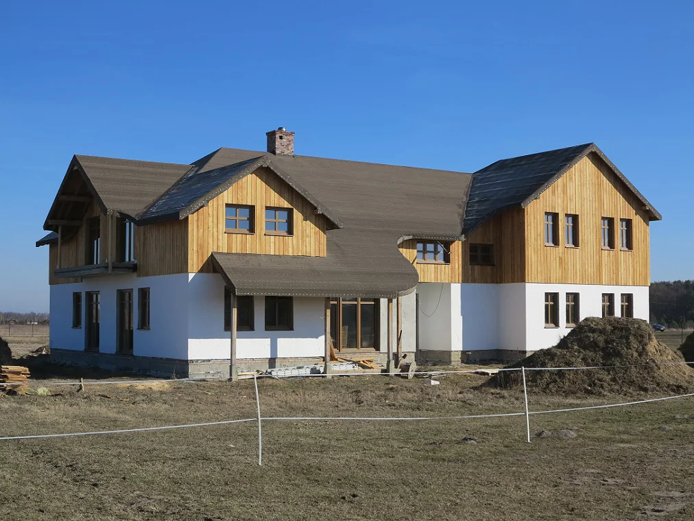 ront i bok domu w trakcie budowy z balkonem; drewniana elewacja, biały tynk i pryzmy ziemi