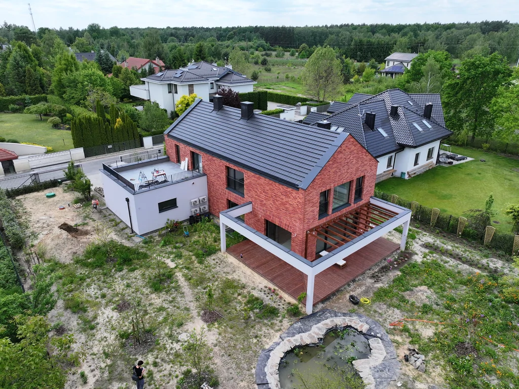 Ujęcie z drona na dom z ceglaną elewacją, tarasem z pergolą oraz ogrodem z oczkiem wodnym