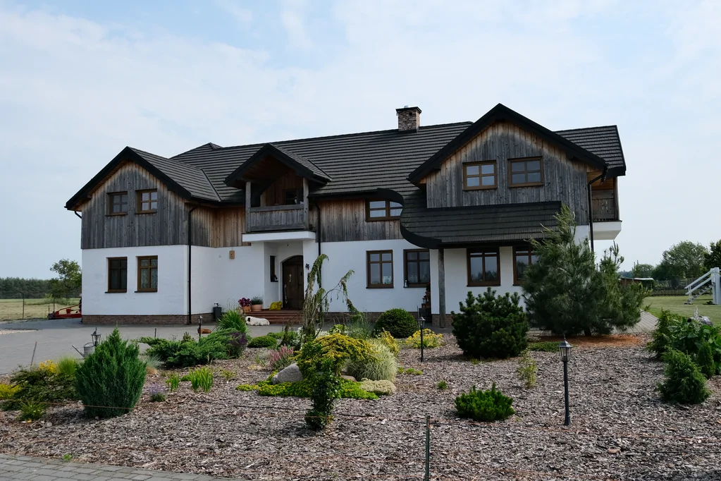 Szeroki widok na front domu z ogrodem: biała elewacja, drewniane poddasze, czarny dach i rabata żwirowa
