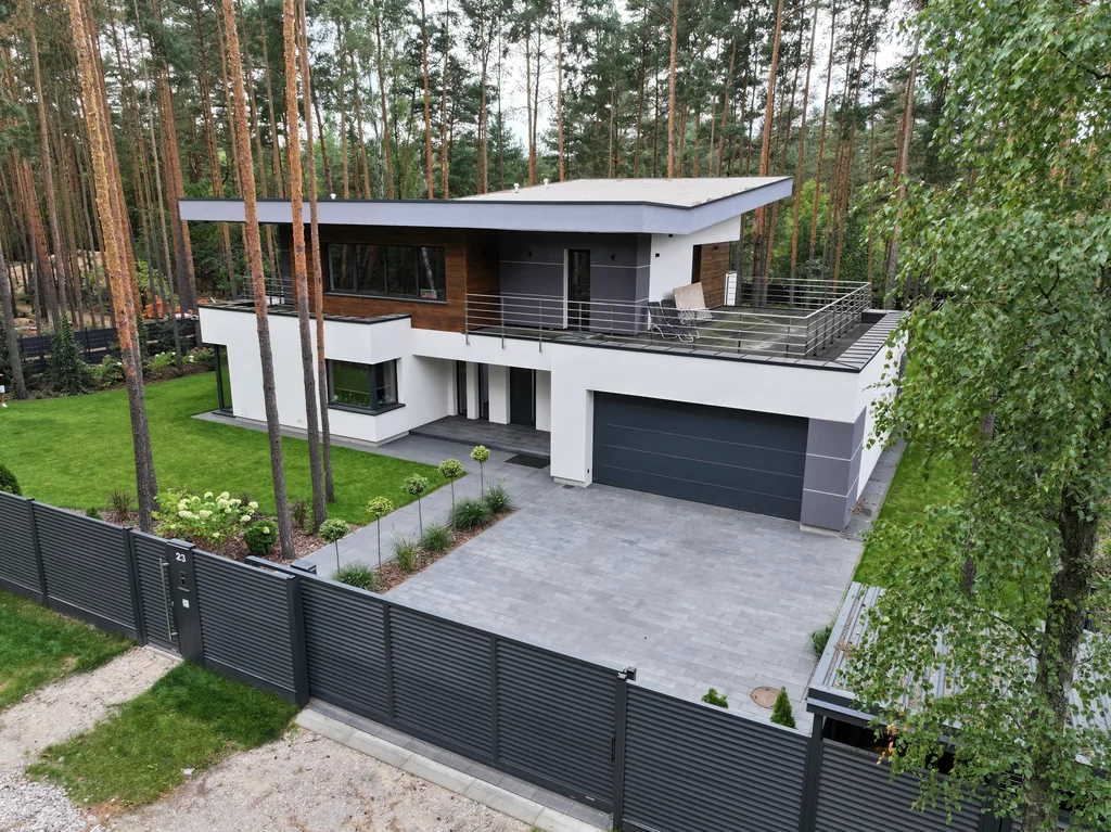 Widok z drona na nowoczesny dom: wysunięte okapy, stalowe balustrady i duży trawnik na leśnej działce