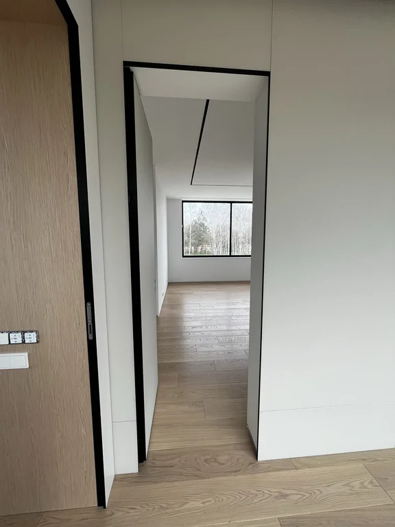 Korytarz z dębową podłogą i ukrytymi drzwiami, prowadzący do pokoju z dużym oknem.