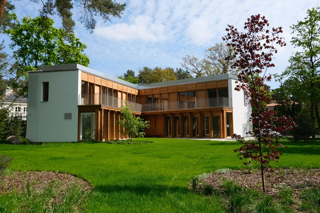 Widok od strony ogrodu na narożną bryłę domu z balkonem, drewnianą elewacją i czerwonym drzewem na pierwszym planie