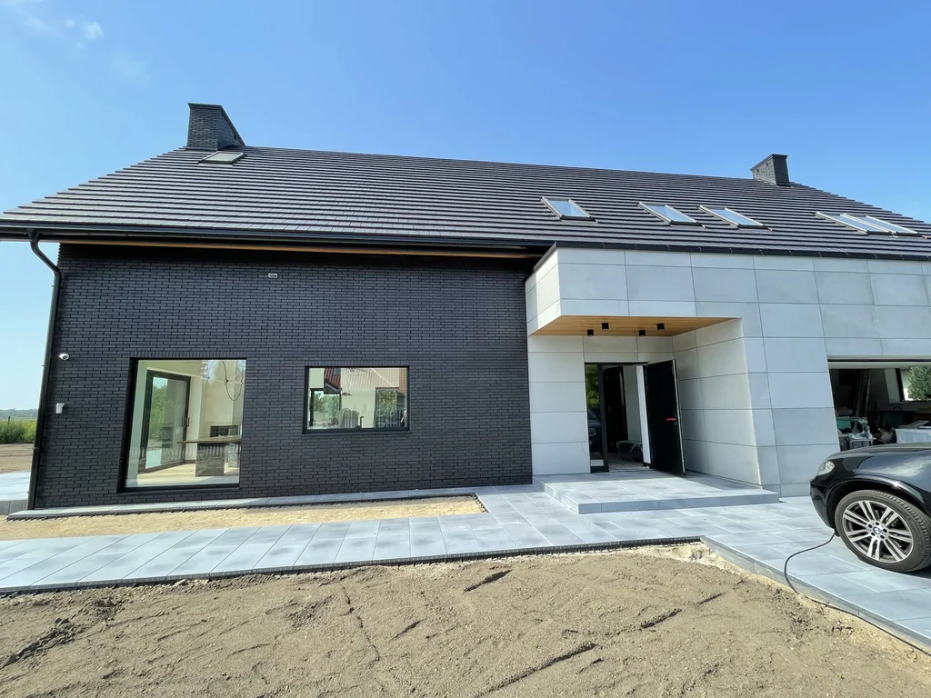 Wejście główne i garaż – jasne płyty elewacyjne, czarna cegła, utwardzony podjazd