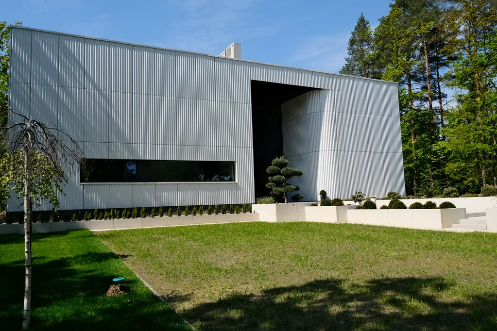 Frontowa elewacja nowoczesnego domu z jasną ryflowaną okładziną, cofniętym wejściem i trawnikiem przed budynkiem