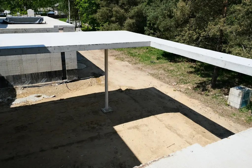 Zadaszony podcień nowoczesnego domu żelbetowego z widoczną płytą stropową i smukłym słupem.