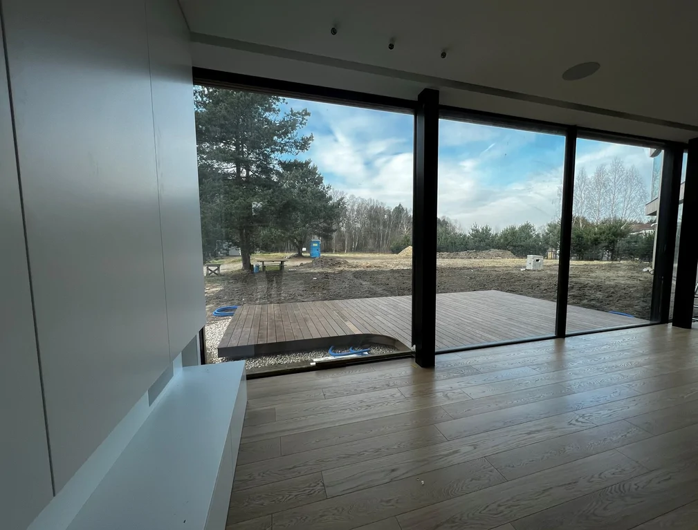 Strefa dzienna z dębową podłogą, zabudową RTV i dużym przeszkleniem na taras z drewnianym deckiem.