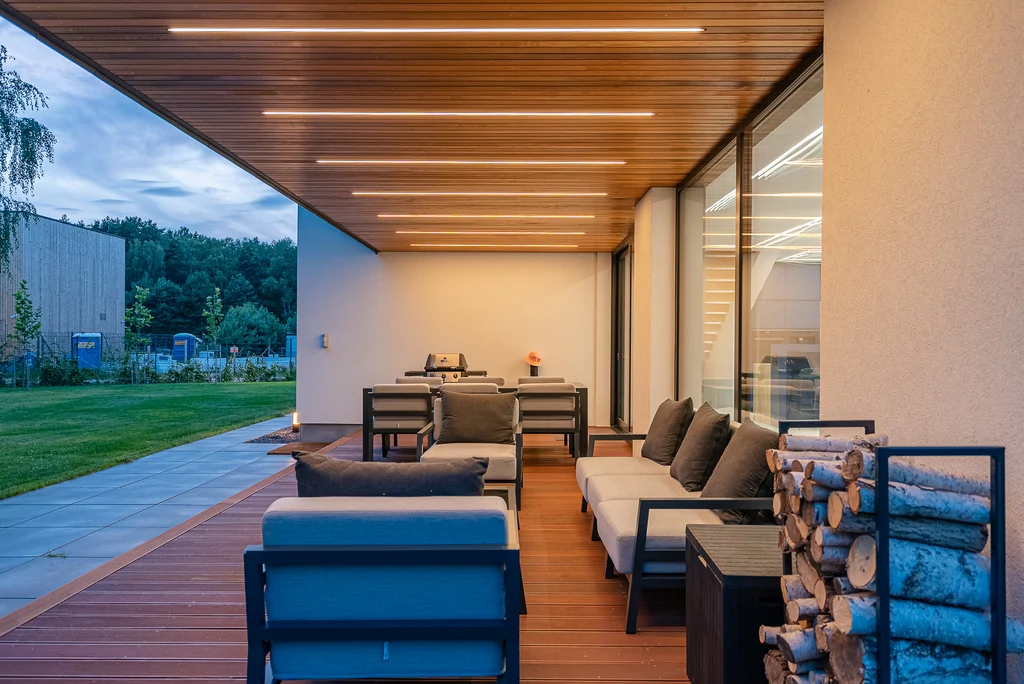 Zadaszony taras z drewnianym sufitem, oświetleniem liniowym i strefą wypoczynku.