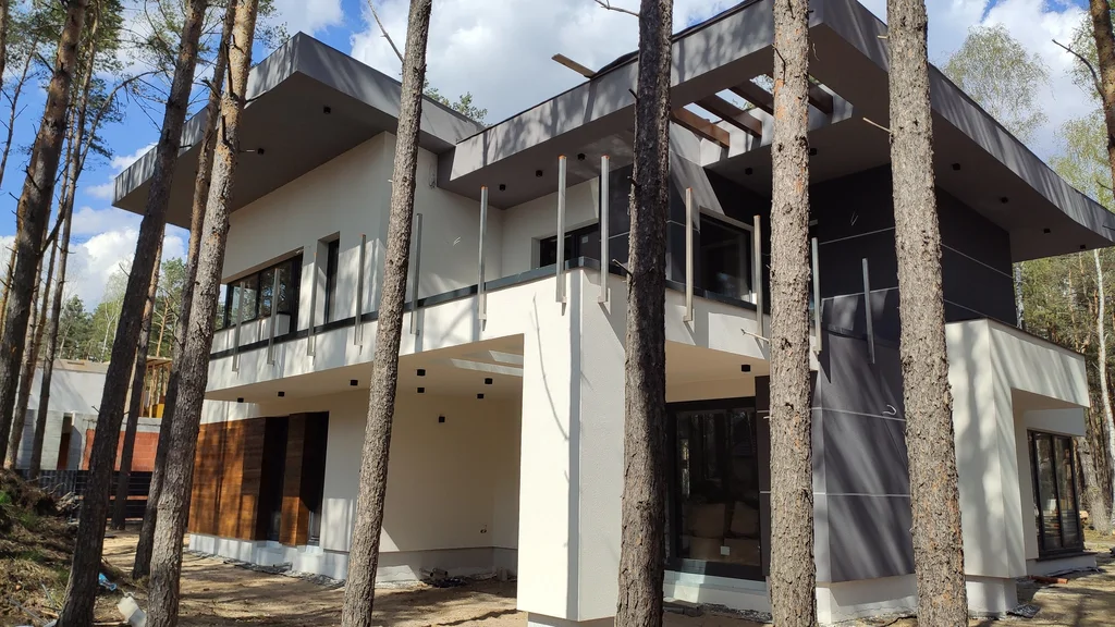 Gotowy dom z tarasem na piętrze i ażurową pergolą w strefie dachu, widok przez sosny