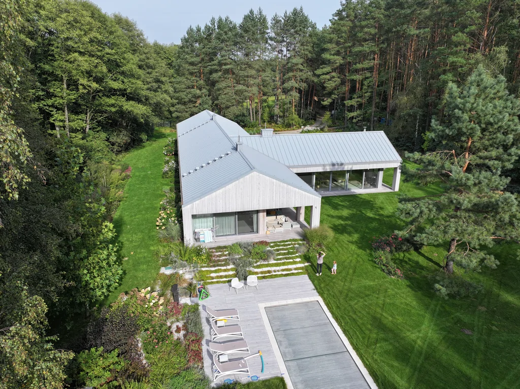 Wysoki ujęcie z lotu ptaka: zabudowa parterowa w kształcie L na tle lasu, ogród z tarasem i basenem (zadaszenie/rola), duże przeszklenia oraz trawniki.