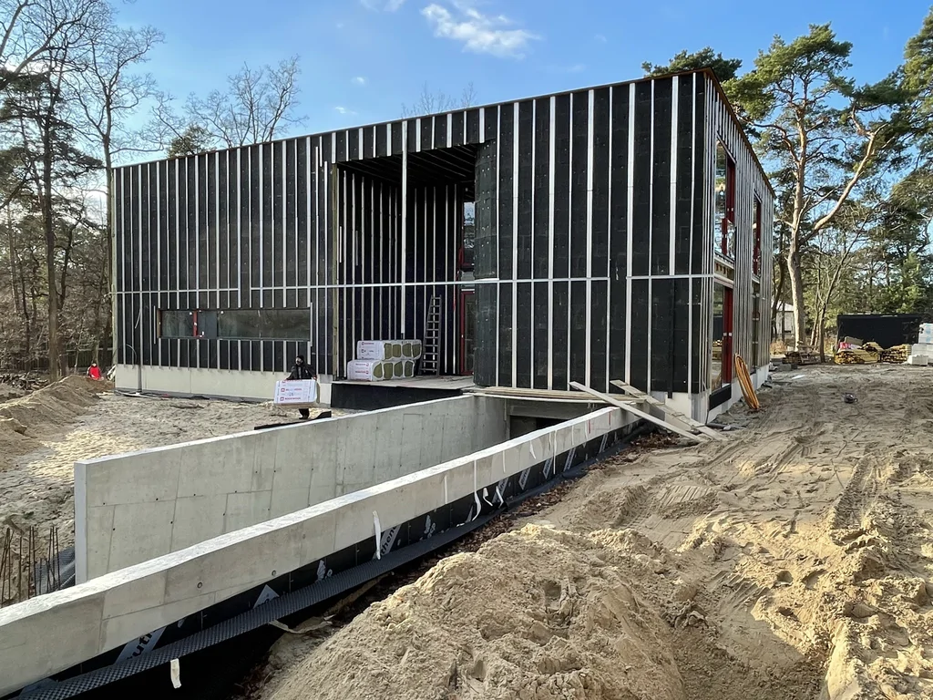 Wejście do nowoczesnego domu z obniżonym dziedzińcem, żelbetową ścianą oporową i ciemną elewacją