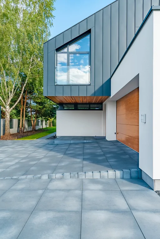 Detal frontowej elewacji domu z nadwieszoną bryłą, wejściem i garażem, ujęcie pionowe.