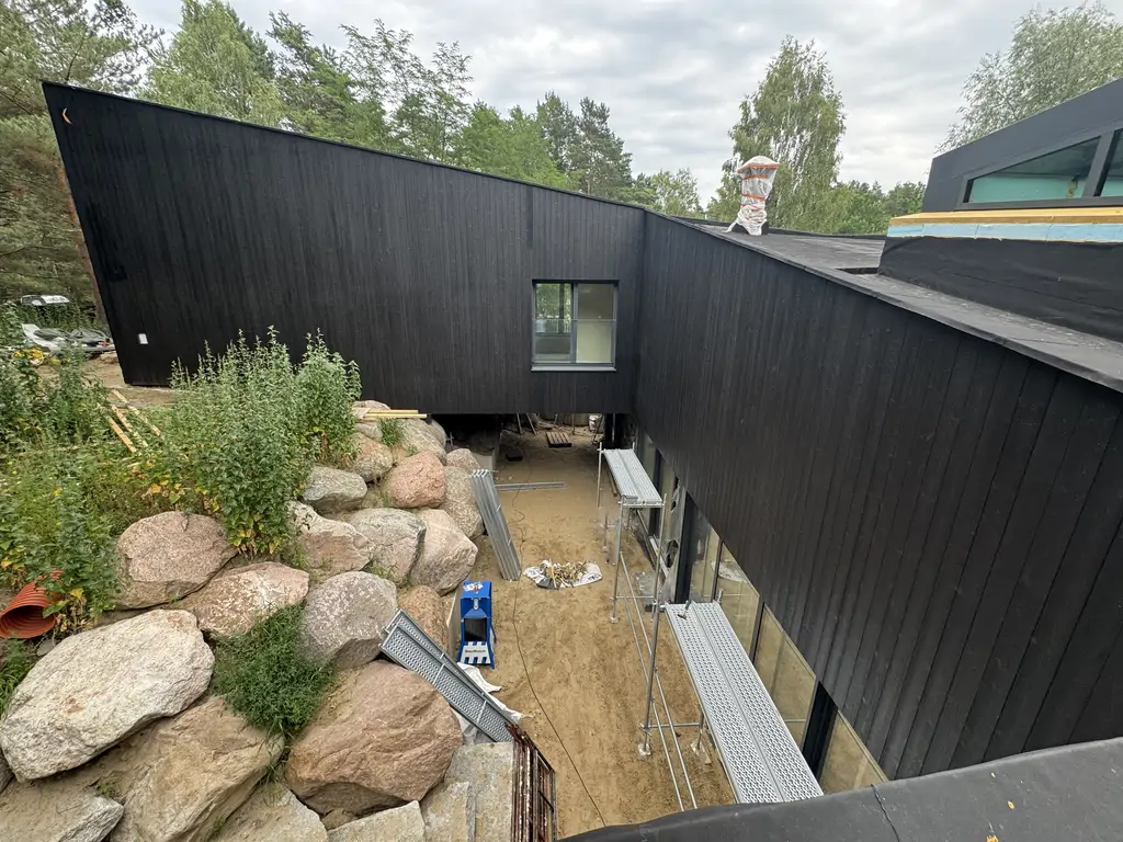 Wewnętrzny dziedziniec domu w trakcie prac, z czarną elewacją, przeszkleniami i skarpą wykończoną dużymi głazami.