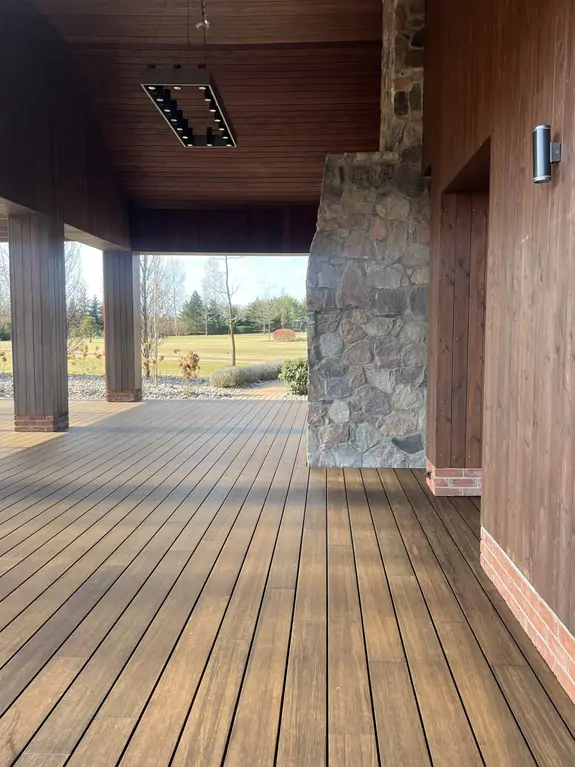 Detal zadaszonego tarasu po remoncie z drewnopodobną nawierzchnią, kamienną ścianą i nowoczesnym oświetleniem sufitowym.