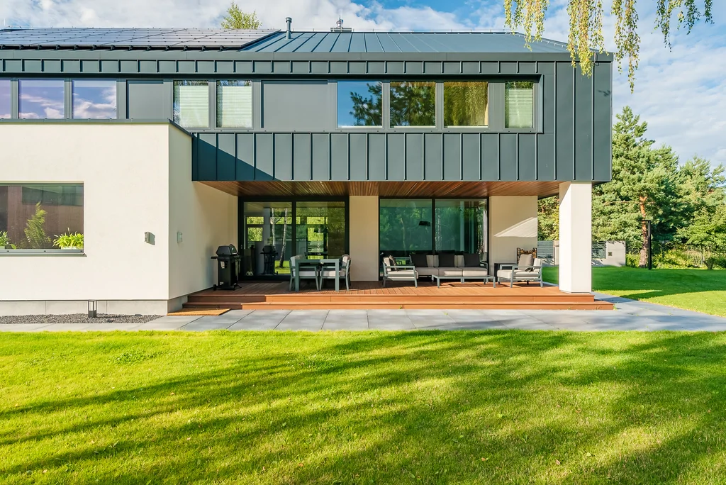 Taras i tylna elewacja domu pod zadaszeniem, z meblami ogrodowymi i dużymi przeszkleniami.