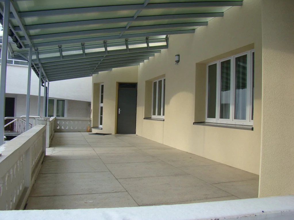 Zadaszony taras przy elewacji; stalowa konstrukcja i posadzka z płyt.