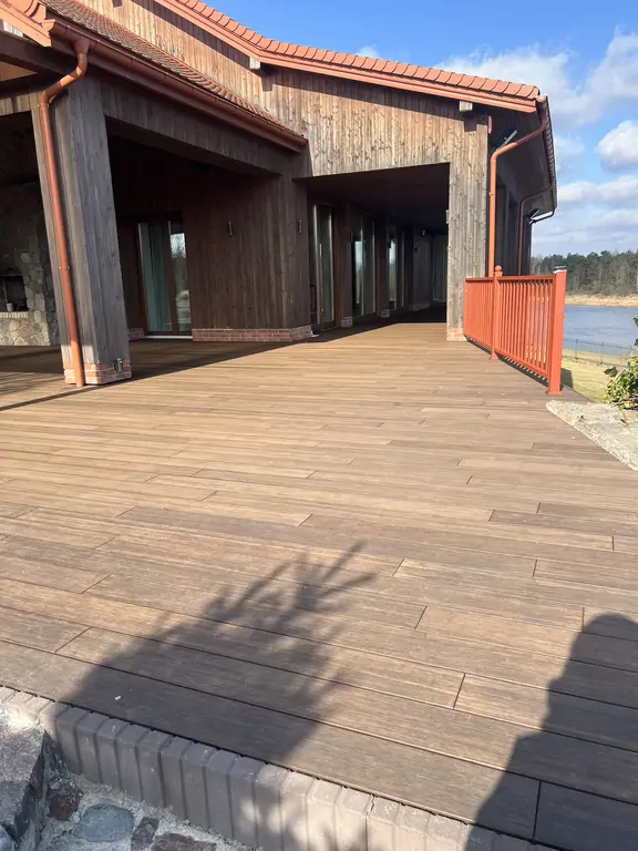 Szeroki taras po remoncie przy domu nad wodą, z drewnopodobną nawierzchnią, czerwonymi balustradami i drewnianą elewacją.