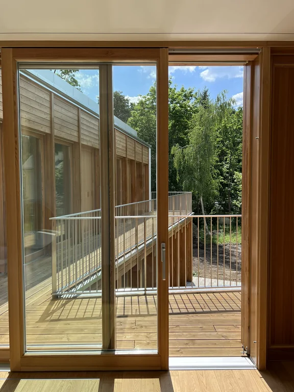 Przeszklone wyjście na drewniany balkon z widokiem na drugie skrzydło budynku i otaczającą zieleń