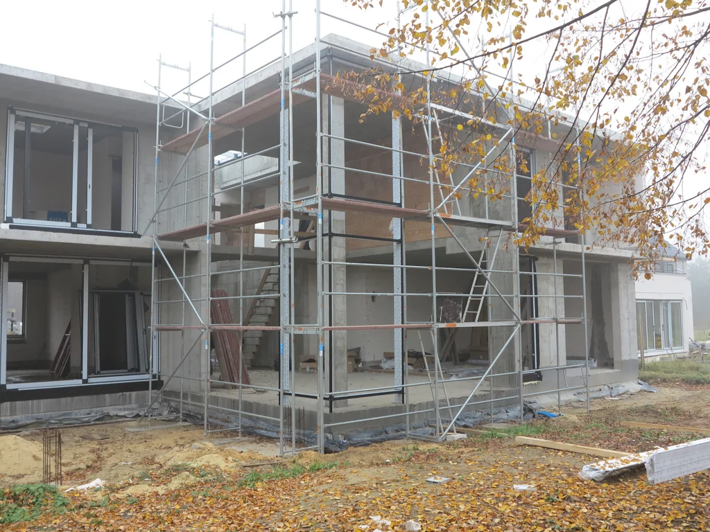 Stan surowy budynku, ściany pustak ceramiczny, dach płaski, montaż stolarki okiennej, widoczne rusztowania 