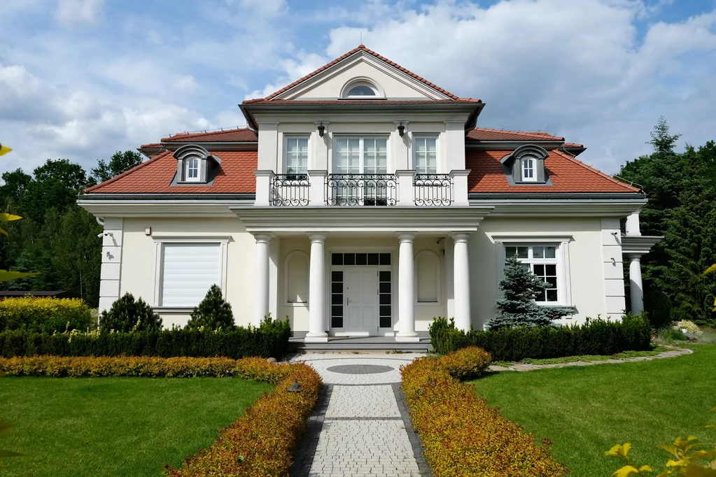 Frontowy widok domu jednorodzinnego z kolumnowym wejściem, balkonem i jasną elewacją, otoczonego przystrzyżonym trawnikiem oraz ozdobnymi krzewami.