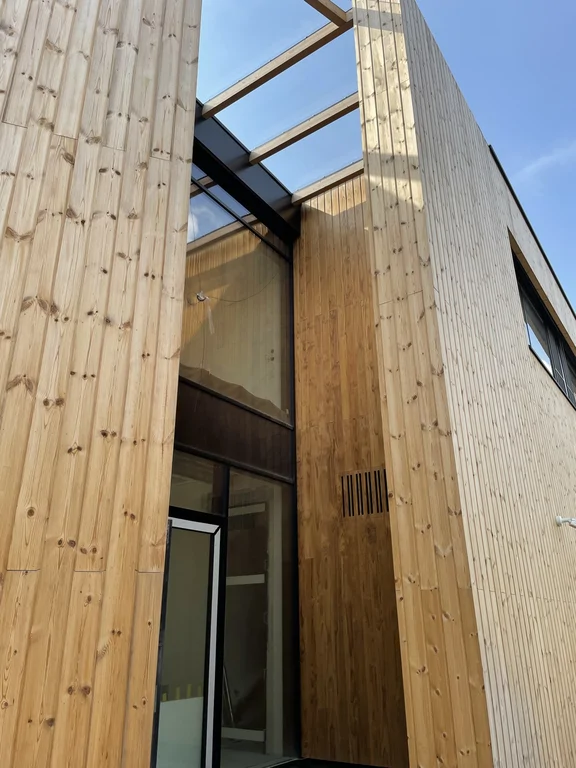 Wnętrze atrium między bryłami: drewniane okładziny, stalowa belka i szklane zadaszenie (świetlik) nad strefą wejściową.