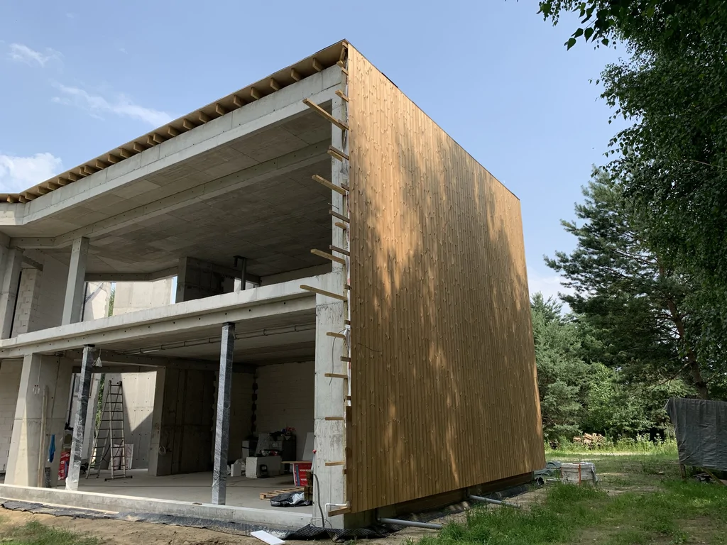 Budynek dwukondygnacyjny w trakcie prac wykończeniowych – widoczna konstrukcja żelbetowa i fragment elewacji z pionowym deskowaniem.