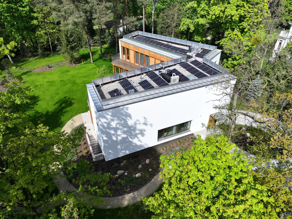 Widok z drona na dach domu z instalacją fotowoltaiczną oraz boczną elewację otoczoną zielenią