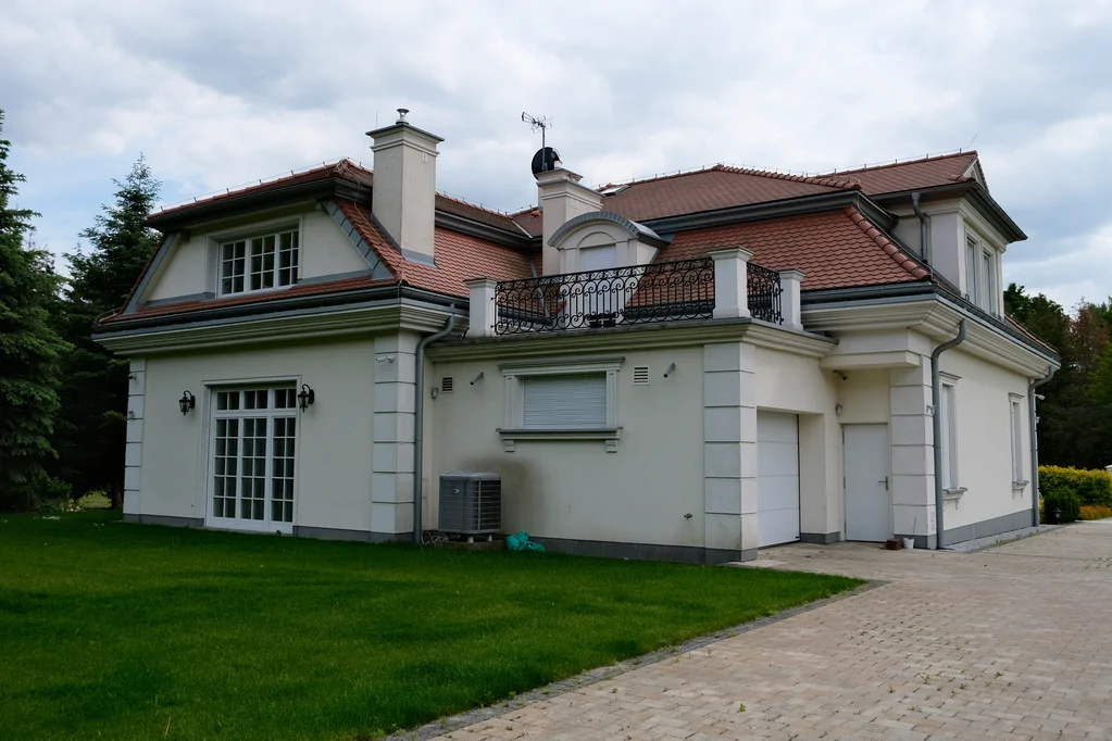 Boczno-tylny widok domu z czerwonym dachem, tarasem na piętrze, dużymi oknami i podjazdem z kostki brukowej, na tle pochmurnego nieba.