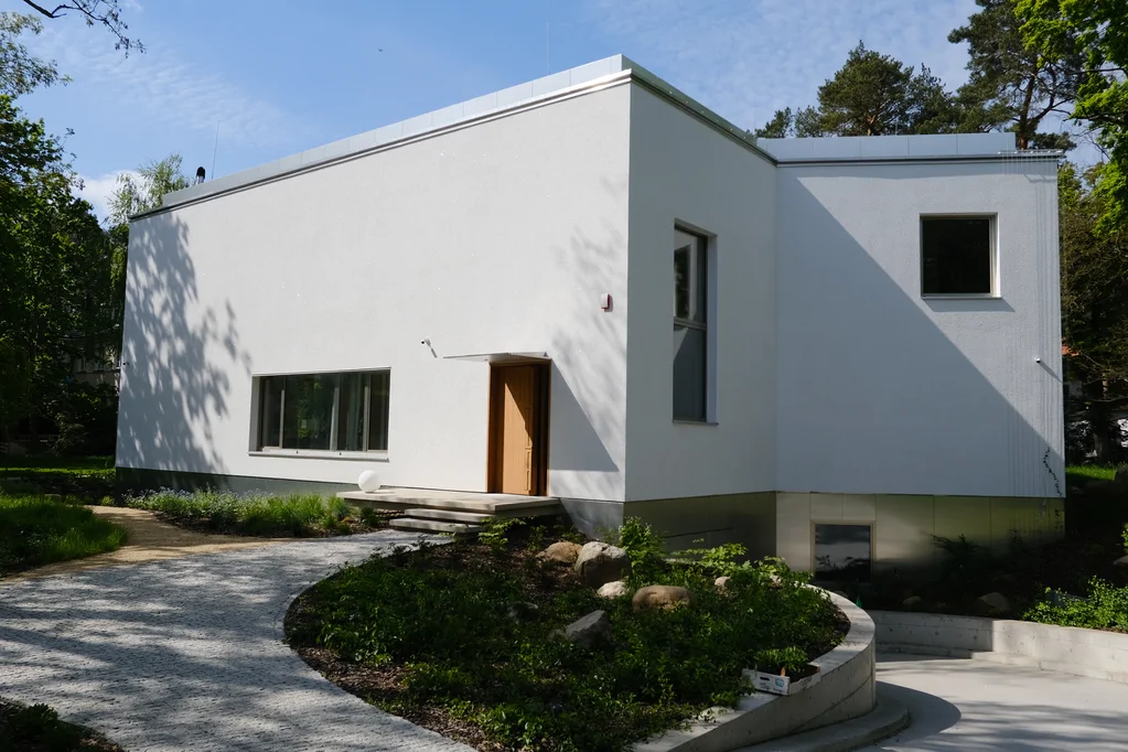 Widok od frontu na białą bryłę domu z wejściem, podjazdem i półkolistą rabatą przy elewacji