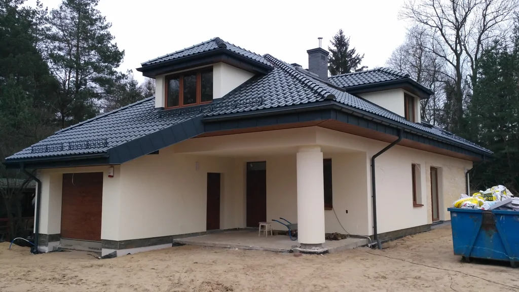 Jasna elewacja strefa wejścia, dach pokryty blachą na rąbek, okrągły słup, brązowa stolarka okien, komin obmurowany klinkierem