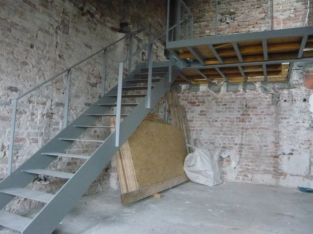 Stalowe schody z poręczą prowadzące na antresolę; surowe ceglane ściany i konstrukcja pod podest.