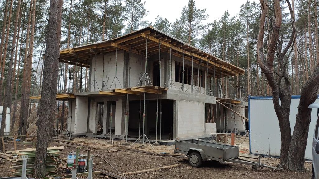 Dom w stanie surowym z podpartym stropem i szalunkami dachu, plac budowy wśród sosen