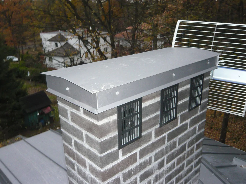 Komin murowany z metalową czapą i kratkami wentylacyjnymi; widok z połaci dachu.