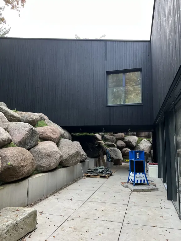 Widok na dziedziniec domu w trakcie realizacji, z czarną elewacją, przeszkleniami i skarpą obłożoną dużymi głazami.