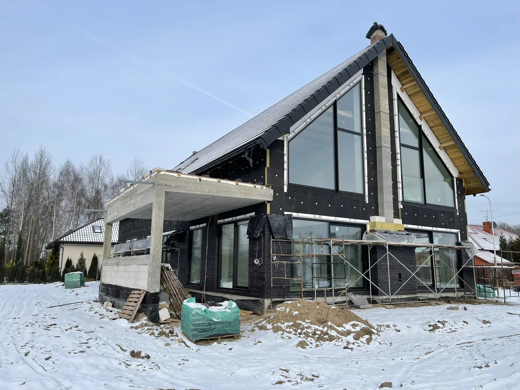 Widok narożny budynku w stanie surowym – dach dwuspadowy, duże przeszklenia i żelbetowy podcień
