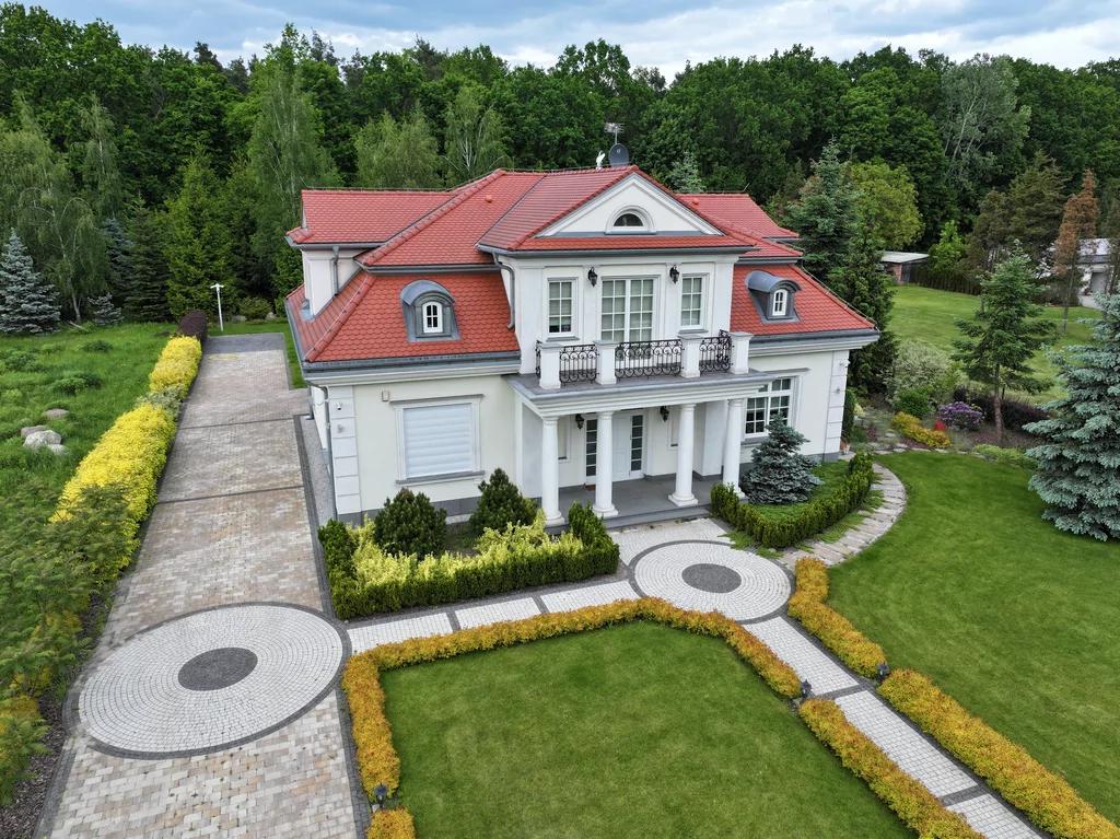 Widok z drona na dom jednorodzinny w stylu dworkowym z białą elewacją z boniami, kolumny okrągłe w strefie wejścia, na balkonie również kolumny i stalowa barierka, pokrycie dachu karpiówka, na dachu widoczne lukarny, przy domu kostka, działka otoczona drzewami 