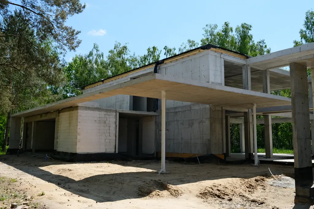 Żelbetowa bryła budynku mieszkalnego w stanie surowym z widocznymi nadwieszeniami i otwartą strefą parteru.