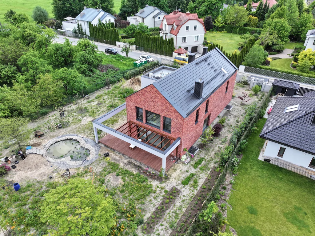 Ujęcie z lotu ptaka pokazujące dom z ceglaną elewacją, pergolą tarasową i zagospodarowaniem działki