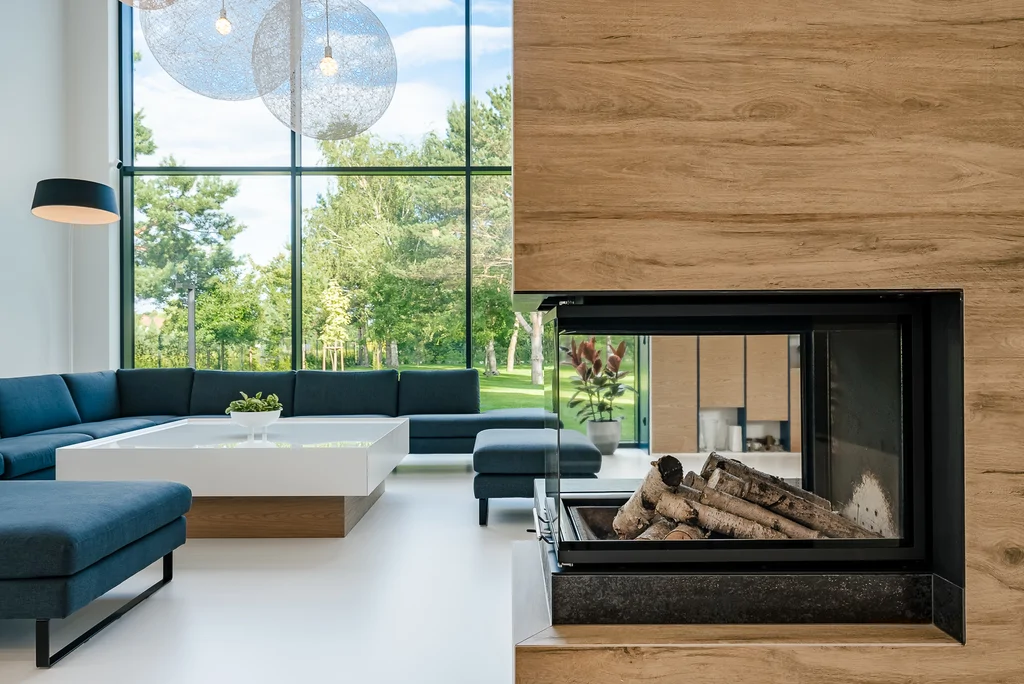Dwustronny kominek w zabudowie drewnopodobnej, z widokiem na salon i ogród.