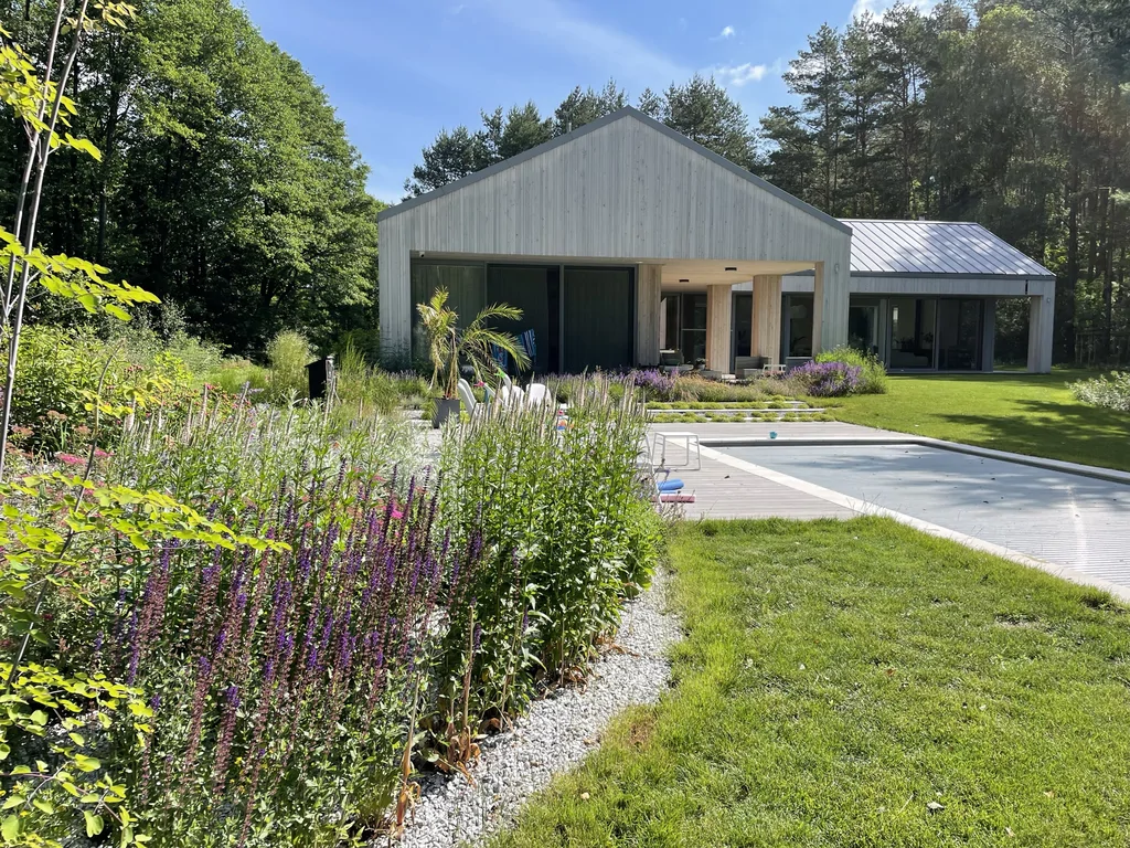 Widok w stronę szczytu budynku: dom z drewnianą elewacją i podcieniem, obok taras i basen z przykryciem; na pierwszym planie rabaty z wysokimi bylinami.