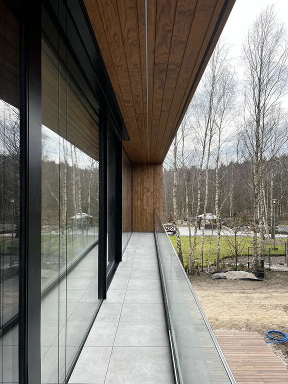 Wąski taras z balustradą szklaną, płytami tarasowymi i drewnianą podbitką. Minimalistyczne detale i mocne kadrowanie widoku.