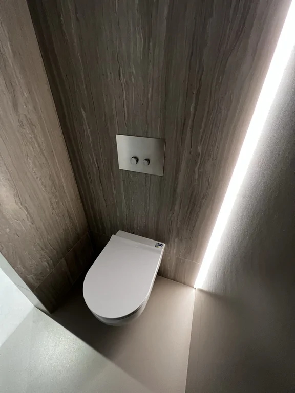 Toaleta z podwieszaną miską WC, okładziną w kamiennym wykończeniu i pionowym podświetleniem wnęki.