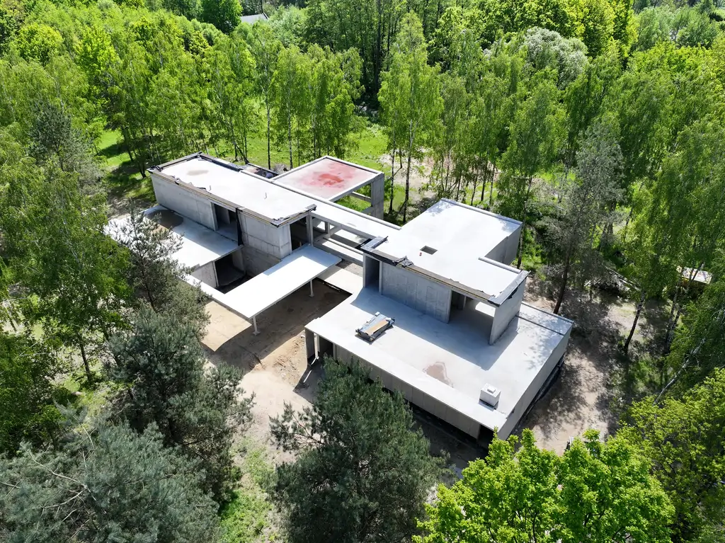 Ujęcie z lotu ptaka przedstawiające bryłę domu żelbetowego w stanie surowym na działce otoczonej drzewami.