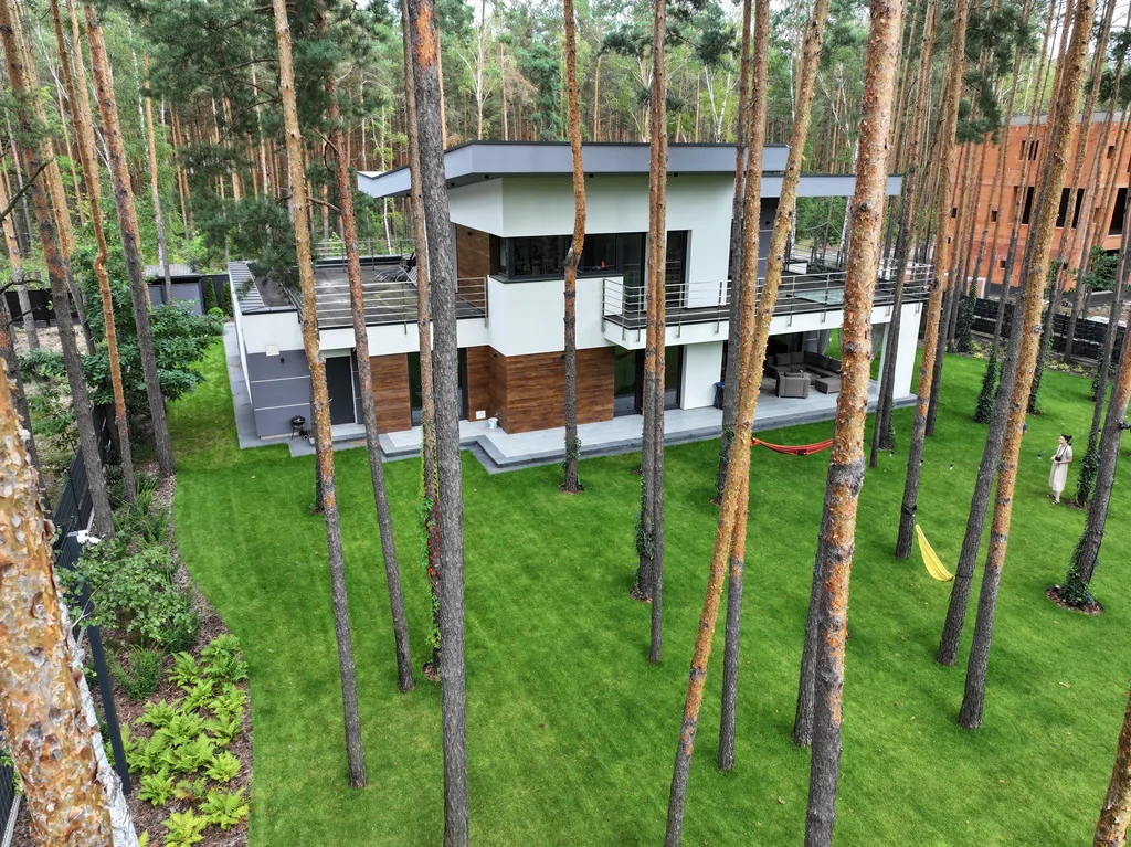 Szeroki kadr z drona: dom w sosnowym lesie z dużym trawnikiem, tarasami i ogrodzeniem wokół działki