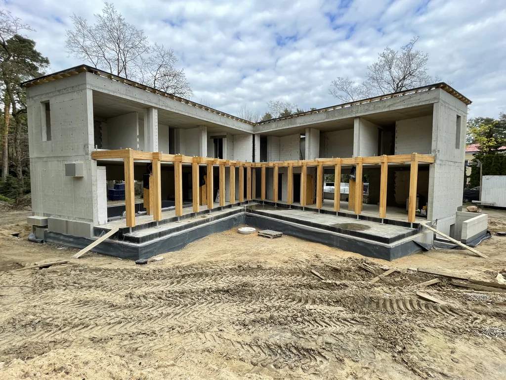 Widok frontowy domu jednorodzinnego w stanie surowym z żelbetową konstrukcją i drewnianą pergolą tarasową