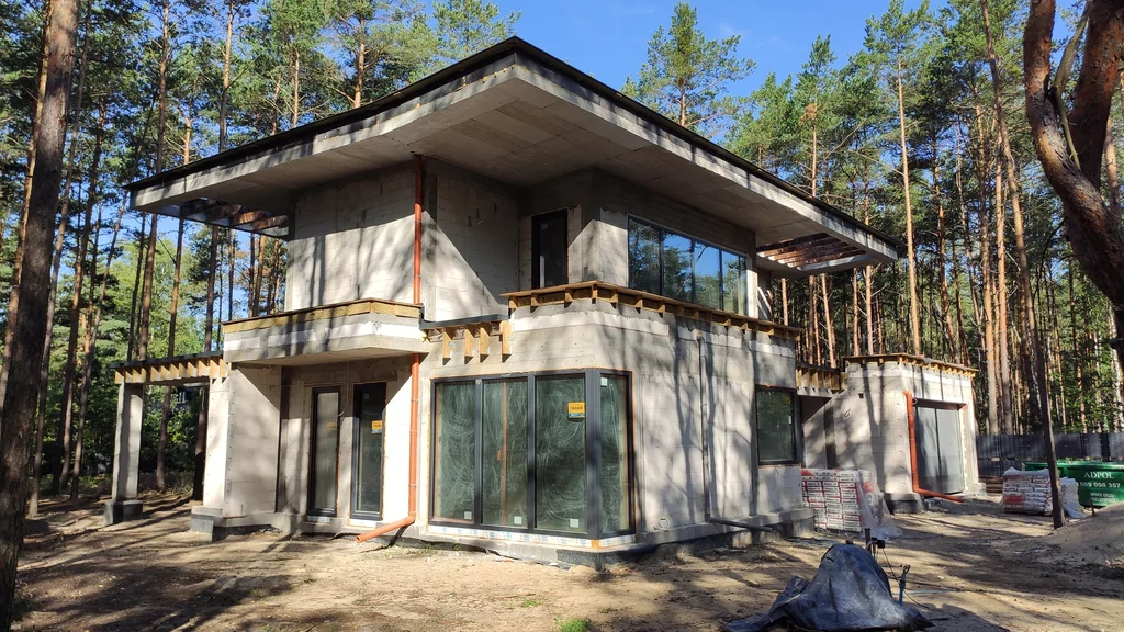 Stan surowy zamknięty: wstawione okna, obróbki dachu i rury spustowe, nowoczesna bryła w lesie