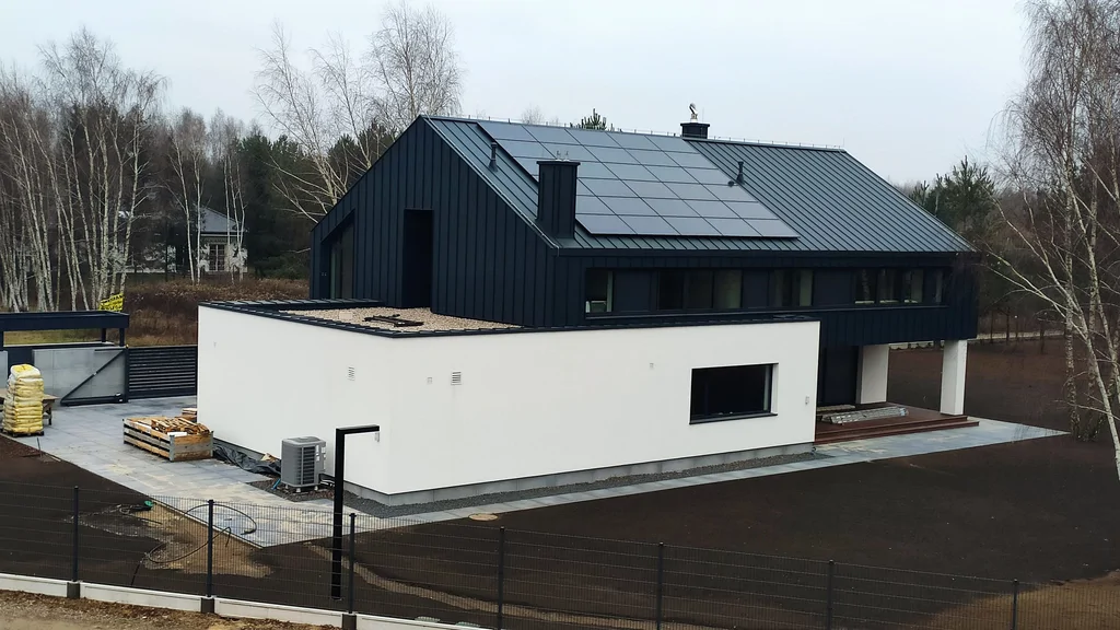 Widok z góry na nowoczesny dom z białą bryłą, czarnym dachem i panelami fotowoltaicznymi