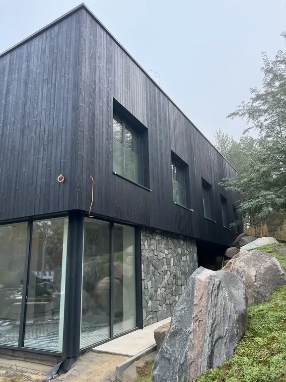 Boczna ściana nowoczesnego domu z czarną elewacją drewnianą, kamiennym detalem i skalną skarpą przy budynku.