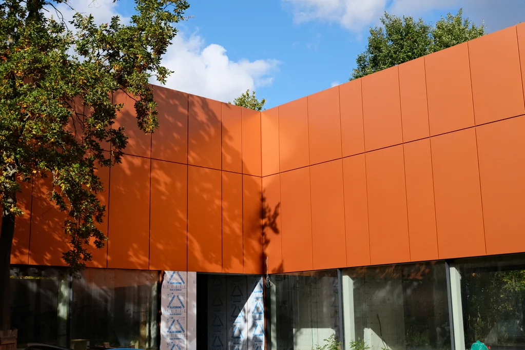 Fragment elewacji corten z cieniem drzewa na tle przeszkleń