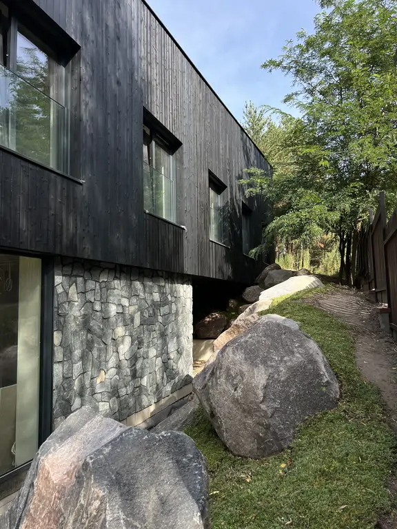 Narożnik nowoczesnego domu z czarną drewnianą elewacją, kamiennym detalem i dużymi oknami od podłogi do sufitu.